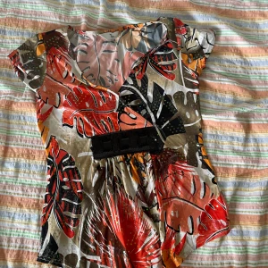 Färgglad blommig topp från Tia - Säljer en superfin och färgglad topp från Tia! Den har ett blommigt mönster i rött, orange, svart och beige. Toppen är kortärmad och har en v-ringad hals. Perfekt för sommarens alla fester eller bara en vanlig dag när du vill känna dig extra fin. Den är i nyskick och har en snygg passform som framhäver midjan med ett svart bälte.