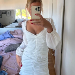 Vit kortklänning från Kiss The Sky - Säljer en superfin vit kortklänning från Kiss The Sky. Klänningen har långa ärmar och en v-ringad hals. Den har en tight passform med volangdetaljer/scrunch där bak och en dragkedja på baksidan💛