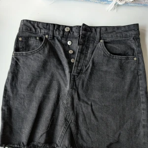 Svart jeanskjol från Perfect Jeans - Säljer en snygg svart jeanskjol från Perfect Jeans. Kjolen har en knappgylf med fyra silverfärgade knappar och två framfickor. Den är kort och har en rå kant nedtill som ger en cool och avslappnad look. Perfekt för både vardag och fest!