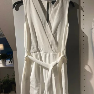 Vit omlottklänning - Säljer en superfin vit playsuit. Den är ärmlös och har ett knytband i midjan som ger en snygg passform. Perfekt för sommarens alla fester eller en fin middag. playsuiten är i nyskick och har en elegant v-ringad design.
