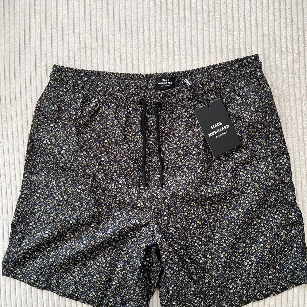 Säljer ett par snygga blommiga shorts från Mads Nørgaard. De är svarta med ett diskret blommönster i vitt och gult. Shortsen har en elastisk midja med dragsko för bästa passform. Perfekta för sommaren! 🌸. Shorts.