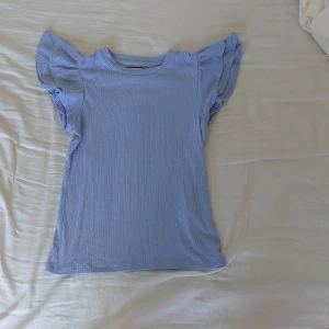 Ljusblå topp från Holly & Whyte - Säljer en söt ljusblå topp från Holly & Whyte by Lindex. Den är i storlek XS och har korta ärmar med volangdetaljer. Är ribbad och gjord av ett mjukt och bekvämt material, perfekt för sommardagar. Den är nästan i nyskick och har en fin passform som är både bekväm och stilren.