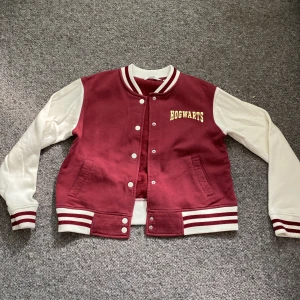 Röd och vit Hogwarts bomberjacka - Säljer en snygg röd och vit bomberjacka med Hogwarts-tema. Jackan har långa vita ärmar och en röd kropp med vita detaljer. På framsidan står det 'Hogwarts' i guld och på baksidan finns Hogwarts vapensköld. Perfekt för alla Harry Potter-fans! Jackan har knappar och ribbade muddar vid ärmslut och nederkant. köparen betalar för frakt 