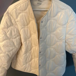 Vit quiltad jacka från MNG - Säljer en snygg vit quiltad jacka från MNG. Jackan har en stilren design med knappar framtill och är perfekt för vår och höst. Den är tillverkad i ett syntetmaterial som gör den lätt och bekväm att bära. Perfekt för att hålla värmen under kyliga dagar! 