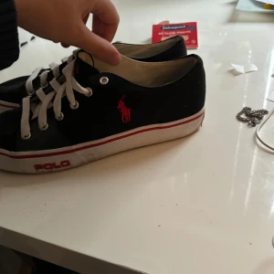 Svarta sneakers från Polo - Säljer ett par svarta sneakers från Polo i bra skick. De har vita snören och en röd logga på sidan. Skorna har en vit sula med en röd rand och texten 'POLO' på hälen. Perfekta för vardagsbruk och ger en stilren look. Pris är inte hugget i sten