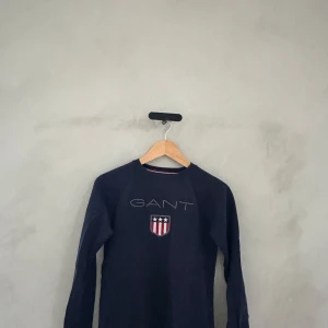 Mörkblå tröja från GANT - Säljer en snygg mörkblå tröja från GANT med deras ikoniska logga och ett coolt emblem på bröstet. Tröjan är långärmad och perfekt för både vardag och lite finare tillfällen. Passar bra till jeans eller chinos för en stilren look.