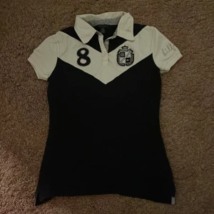 Svart och vit pikétröja från Tommy Hilfiger - Säljer en snygg svart och vit pikétröja från Tommy Hilfiger i storlek XS. Tröjan har korta ärmar och ett coolt emblem på bröstet samt siffran 8. pris kan diskuteras💕