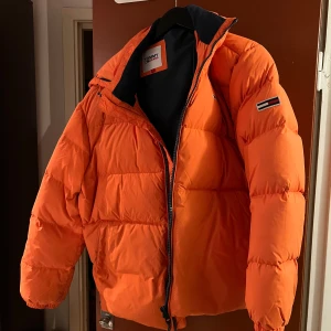 Orange pufferjacka från Tommy Hilfiger - Säljer en snygg orange pufferjacka från Tommy Hilfiger. Den har en dragkedja framtill och en mysig huva med snörning. Perfekt för kalla dagar och ger en cool look med sin starka färg. Jackan är i mycket bra skick!