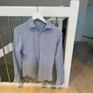 Blå skjorta från Xacus - Säljer en stilren blå skjorta från Xacus i storlek M. Skjortan är långärmad och har klassiska knappar framtill. Perfekt för både vardag och festliga tillfällen. Materialet känns lätt och bekvämt, vilket gör den idealisk för vår och sommar.