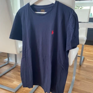 Mörkblå t-shirt från Ralph Lauren - Säljer en snygg mörkblå t-shirt från Ralph Lauren i storlek M. Den har en klassisk design med korta ärmar och en liten röd broderad logga på bröstet. Perfekt för en avslappnad stil eller sommarens äventyr. T-shirten är i bra skick och gjord av mjuk bomull.