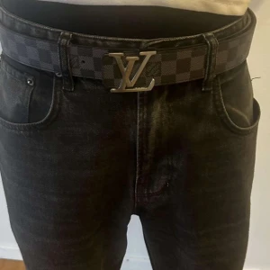 Rutig Levi Vitteon bälte - Säljer ett snyggt bälte i rutig design med en stilren spänne i silver. Bältet är gjort av skinn och har en klassisk svart och grå färgkombination. Perfekt för att ge din outfit en lyxig touch. Passar både till vardags och fest!
