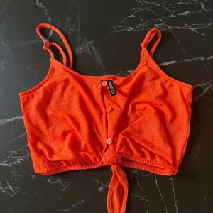 Orange croppad linnetopp från Cubus - Snygg orange croppad linnetopp från Cubus med knappar framtill och knytning i nederkant. Perfekt för sommardagar eller en avslappnad fest. Den är ärmlös och har justerbara axelband för bästa passform. En färgglad och trendig topp att ha i garderoben!