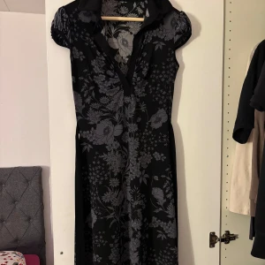 Svart blommig klänning - Snygg svart klänning med blommigt mönster i grått. Den har korta ärmar och en klassisk krage, perfekt för både vardag och fest. Klänningen är i ett mjukt och bekvämt material som faller fint. Storlek s/m
