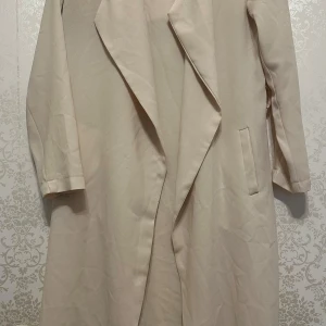 Beige trenchcoat - Säljer en stilren beige trenchcoat i normal passform. Perfekt för vår och höst med långa ärmar och klassisk design. Den har en elegant krage och är gjord i ett lätt material som passar till alla tillfällen.
