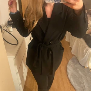 Svart kappa med bälte - Säljer en svart kappa med ett band i midjan från Zara.  Den har långa ärmar och knappar framtill. Perfekt för höst och vår. Säljer för jag får ingen användning av den då jag har en annan kappa!