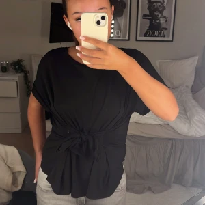 Svart topp med knytband - Säljer en svart topp med korta ärmar och knytband i midjan.Toppen har en lös passform och är superbekväm. Passar bra till jeans eller kjol.