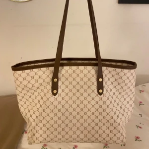 Beige och brun shoppingväska - Säljer en super snygg off white/beige shoppingväska med brunt mönster och detaljer. Väskan har långa handtag och en dragkedja upptill. Perfekt för shoppingturer eller som en stilren vardagsväska. Den är rymlig och har en innerficka med dragkedja för extra säkerhet.