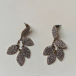 Glittriga örhängen i bladform - Säljer ett par superfina örhängen i bladform med massor av glittriga stenar. De är i en elegant guldton och mäter cirka 7 cm i längd. Perfekta för att lysa upp vilken fest som helst! ✨