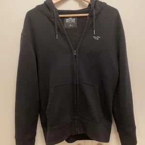 Svart hoodie från Hollister - Säljer en svart hoodie från Hollister Must-Have Collection i storlek XS. Den har en dragkedja framtill och en broderad logga på bröstet. Perfekt för en avslappnad stil och passar till alla säsonger. Supermjuk och bekväm!