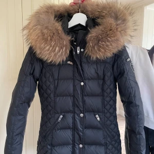 Svart dunjacka från Rockandblue - Säljer en snygg svart dunjacka från Rockandblue i storlek 34. Jackan har en stor, fluffig pälsfodrad huva och quiltad design. Säljer då jag växt ur den! Använt fåtal gånger, inga defekter 