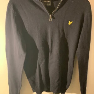 Mörkblå tröja med dragkedja från Lyle & Scott - Snygg mörkblå tröja från Lyle & Scott med en liten gul logotyp på bröstet. Tröjan har en dragkedja vid halsen och långa ärmar. Perfekt för en stilren look.