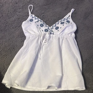 Vit ärmlös topp med broderi - Säljer en söt vit ärmlös topp med blått broderi och smala axelband. Toppen har en v-ringad design och knytband i midjan för en justerbar passform. Perfekt för varma sommardagar!