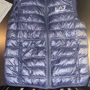 Blå dunväst från Emporio Armani - Snygg blå dunväst från Emporio Armani med EA7-logga på bröstet. Västen har en quiltad design och dragkedja framtill. Perfekt för lager-på-lager under kyligare dagar.
