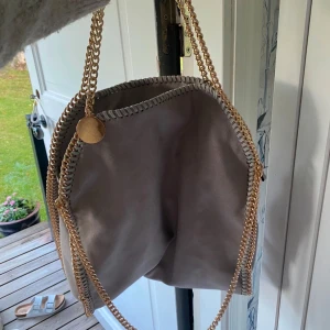 Beige axelväska med kedjedetaljer - Snygg beige Stella McCartney liknande axelväska med guldfärgade kedjor som axelremmar och dekorativa detaljer längs kanterna. Väskan har en mjuk och stilren design, perfekt till skolan eller jobbet. Endast använd 1 gång 🤍