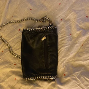 Svart axelväska med kedjedetaljer från Tiamo - Snygg svart axelväska från Tiamo med coola kedjedetaljer runt kanterna och axelremmen. Väskan har dragkedja på baksida för extra förvaring. Knappt använd. Priset kan diskuteras. 