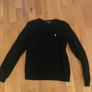 Svart stickad tröja från Ralph Lauren Sport - Säljer en stilren svart stickad tröja från Ralph Lauren Sport. Tröjan har ett diskret kabelstickat mönster och en broderad logotyp på bröstet. Perfekt för kyligare dagar med sin mjuka bomullskvalitet.