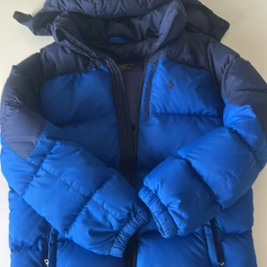 Blå dunjacka från Polo Ralph Lauren - Säljer en snygg blå dunjacka från Polo Ralph Lauren. Jackan har en broderad logga på bröstet och dragkedja framtill. Perfekt för kyliga dagar med sin vadderade design och praktiska fickor.
