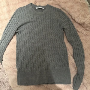 Grå stickad tröja från Zara - Säljer en stilren grå stickad tröja från Zara. Tröjan har ett klassiskt flätmönster och långa ärmar, perfekt för kyligare dagar. Den är mjuk och bekväm, idealisk för både vardag och avslappnade tillfällen. Den är även tajt.