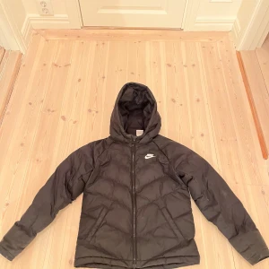 Svart dunjacka från Nike - Säljer en svart dunjacka från Nike med huva och dragkedja. Perfekt för kyliga dagar med sin stilrena design och varma material. Jackan har långa ärmar och en diskret Nike-logga på bröstet.