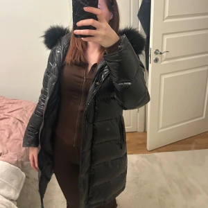 Svart dunjacka med pälshuva - Snygg svart dunjacka med en fluffig pälshuva. Jackan har en lång design och stängs med dragkedja. Perfekt för kalla dagar.