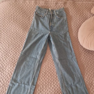 Blå jeans från Weekday - Säljer ett par klassiska blå jeans från Weekday i modellen Rowe. De har en hög midja och raka ben med en dragkedja och knapp framtill. Perfekta för en avslappnad stil.