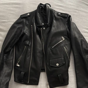 Svart skinnjacka med dragkedjor - Snygg svart skinnjacka med flera dragkedjor och knappar. Jackan har en klassisk bikerstil med axelklaffar och asymmetrisk dragkedja framtill. Perfekt för en cool och edgy look.