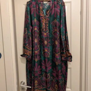 Mönstrad långklänning i grönt och rosa - Säljer en färgglad långklänning med ett unikt mönster i grönt och rosa. Klänningen har långa ärmar och en lös passform, perfekt för en bohemisk stil. Den har en v-ringad hals och knappar framtill för en avslappnad look. NYSKICK storlek M/L