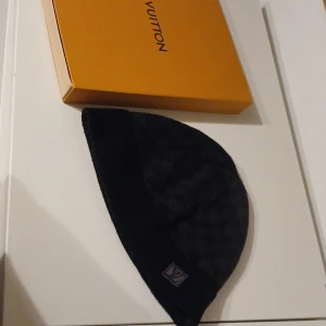 Svart mössa från Louis Vuitton - Snygg svart mössa från Louis Vuitton med diskret rutigt mönster. Mössan har en broderad logotyp framtill och är tillverkad i ett mjukt material. Perfekt för att hålla stilen under kyliga dagar. Fått i present av en vän har inget kvitto bara lådan knappt använd