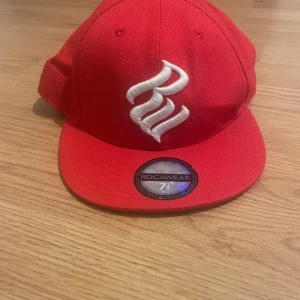 Röd keps från Rocawear - Snygg röd keps från Rocawear med broderad logga framtill och text baktill. Tillverkad i en blandning av akryl och ull för en bekväm passform. Perfekt för att ge din outfit en cool touch.