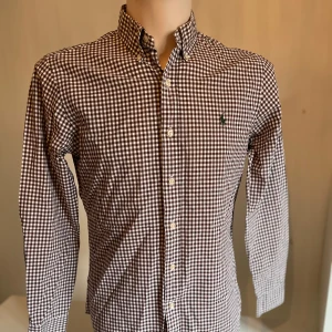 Brun och vit rutig skjorta från Ralph Lauren - Snygg brun och vit rutig skjorta från Ralph Lauren med klassisk krage och knappar framtill. Skjortan har långa ärmar och en liten broderad logotyp på bröstet. Perfekt för en stilren look!