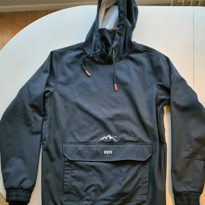 Svart vinterjacka från Dope - Säljer en svart vinterjacka från Dope med huva och stor ficka framtill. Jackan har långa ärmar och en cool logga på bröstet. Materialet är en blandning av nylon och spandex vilket gör den både bekväm och stretchig. Perfekt för kyliga dagar! ❄️
