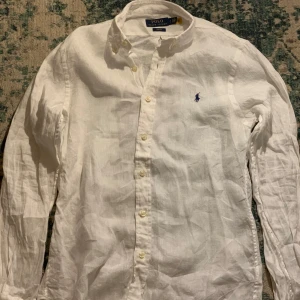 Vit linneskjorta från Polo Ralph Lauren - (ORD PRIS- 2100)Snygg vit linneskjorta från Polo Ralph Lauren i slim fit. Skjortan har långa ärmar och klassiska knappar framtill. Den är tillverkad i ett lätt och bekvämt material, perfekt för en stilren look. Den ikoniska loggan är broderad på bröstet.