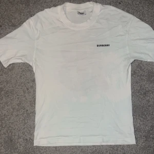 Vit t-shirt från Burberry - Vit t-shirt från Burberry med ett stort, mönstrat TB-tryck på ryggen och en liten logga på bröstet. Perfekt för en stilren och lyxig look.