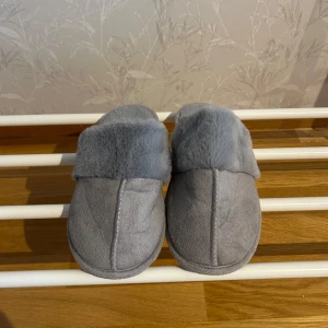 Grå tofflor med fuskpäls - Mysiga grå tofflor med mjuk fuskpäls på insidan och utsidan. Perfekta för att hålla fötterna varma under kalla dagar. Sulan är i en matchande grå färg och storleken är 37/38.