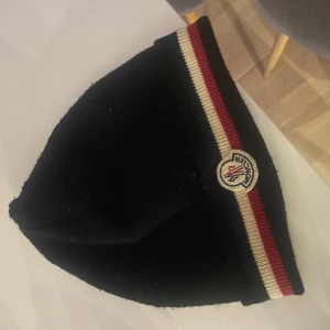 Svart mössa från Moncler - Snygg svart mössa från Moncler med en röd och vit randdetalj. Den har en broderad Moncler-logga framtill. Perfekt för att hålla värmen med stil.