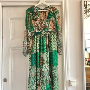 Grön mönstrad klänning från Zara - Säljer en vacker grön mönstrad klänning från Zara med volangdetaljer och v-ringning. Klänningen har långa ärmar och ett bohemiskt mönster i olika gröna nyanser. Perfekt för en somrig look!