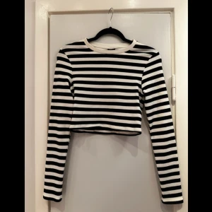 Randig croppad tröja från Gina Tricot - Säljer en svartvit randig croppad tröja från Gina Tricot. Tröjan är tillverkad i mjuk bomull med en liten andel elastan för stretch. Den har långa ärmar och en klassisk rund halsringning. Perfekt för en stilren look!  Mått:  Byst: 36 cm Midja: 35 cm Längd: 37 cm Innerärm: 50 cm