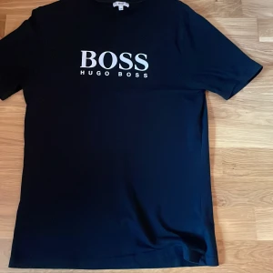Svart t-shirt från Hugo Boss - Snygg svart t-shirt från Hugo Boss med vit logga på bröstet. Perfekt för en stilren look. Kortärmad och tillverkad i mjukt material för bekväm passform.