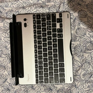 Silverfärgat tangentbord - Säljer ett silverfärgat tangentbord med svarta tangenter. Det har en kompakt design och är perfekt för att kopplas till en surfplatta. Tangentbordet har en hållare för att enkelt fästa en enhet och ger en smidig skrivupplevelse.