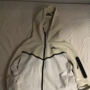 Vit hoodie från Nike - Säljer en stilren vit hoodie från Nike med dragkedja och huva. Jackan har svarta detaljer och en ficka på ärmen. Perfekt för en sportig look.